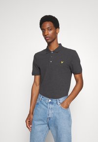Donkergrijze polo met korte mouwen, drie knopen bij de hals en een klein geel logo op de linkerborst, gecombineerd met lichtblauwe spijkerbroek.