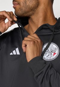 Schwarzes Hoodie mit einem Reißverschlusskragen, das ein kreisförmiges Logo und drei weiße Streifen auf der Schulter hat. Glatte Textur, athletische Passform.