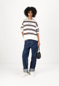Valge sviiter mitmevärviliste triipudega, bränditud "GUESS JEANS". Koos vabade siniste teksade ja valgete tossudega, täiendab must kott.