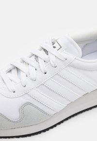 Bílé tenisky Adidas s designem tří pruhů, se šněrováním, se semišovým akcentem poblíž podešve a černou podešví s drážkami.