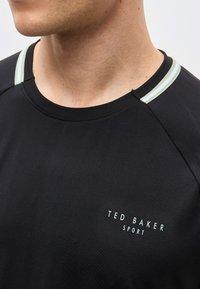 Κοντινό πλάνο του λαιμού και του πάνω μέρους του στήθους ενός ατόμου που φοράει μαύρο t-shirt Ted Baker Sport με ανοιχτόχρωμες ρίγες στις ραφές των ώμων.