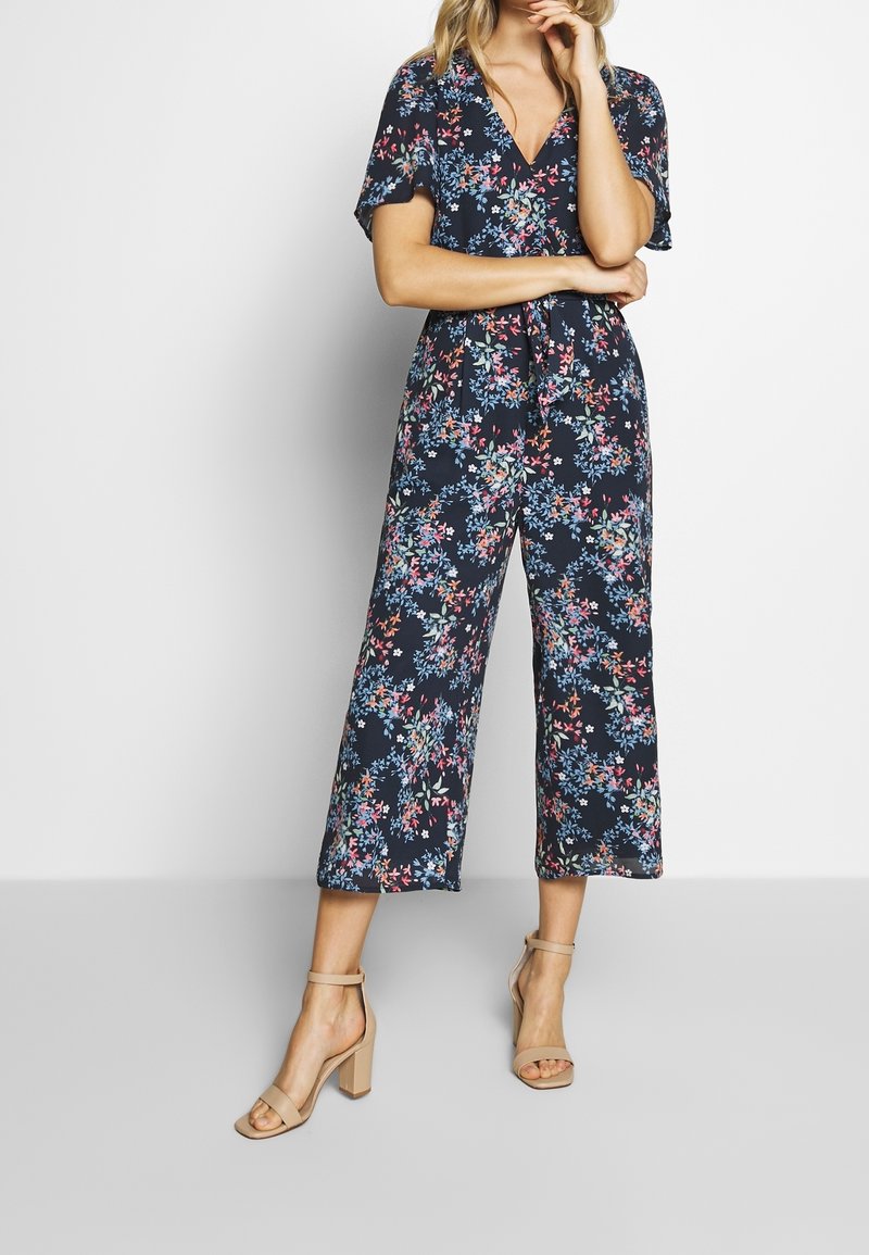 Kombinezon w kwiaty na granatowym tle, z dekoltem w kształcie litery V, krótkimi rękawami i szerokimi nogawkami. Posiada wielokolorowe wzory kwiatowe.