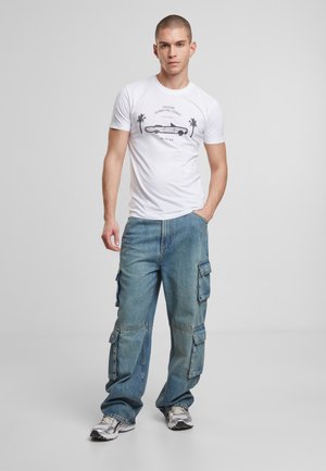 Tricou alb din bumbac cu un grafic al unei mașini și palmieri. Pantaloni cargo de culoare albastru deschis, cu o croială lejeră și buzunare laterale. Pantofi sport gri.