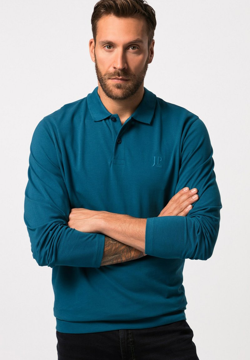 Teal pitkähihainen polo-paita, jossa on klassinen kaulus, kolmesta napista koostuva nappilista ja kirjailtu logo rinnassa, yhdistettynä tummiin farkkuihin.