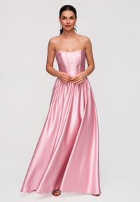 Femme portant une robe bustier en satin rose, avec un corsage ajusté et une jupe fluide, debout devant un fond clair uni.