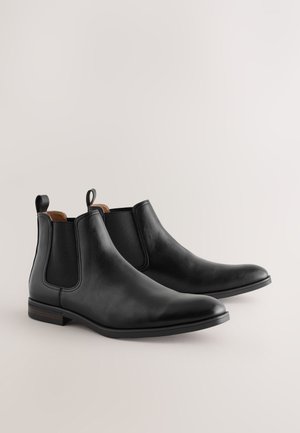 Schwarze Chelsea-Stiefel aus Leder mit einer schlanken, spitzen Zehenpartie, elastischen Seitenteilen und einem braunen Lederakzent am Schaftabschluss. Gummisohlen.