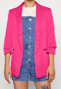 Blazer rosa acceso con colletto a punta, maniche a tre quarti e tasche frontali, indossato sopra una salopette di jeans con bottoni argentati.
