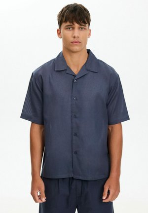 Joven con cabello corto y oscuro, vestido con una camisa de manga corta azul marino abotonada y pantalones a juego sobre un fondo liso.