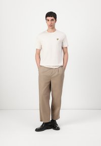 T-shirt crème à manches courtes avec un logo jaune, associé à un pantalon large beige et des chaussures noires, se tenant devant un fond uni.