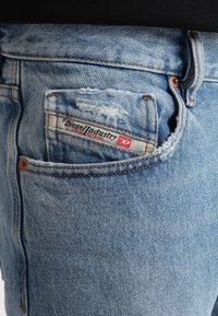 Ljusblå slitna denimjeans med en bakficka som har en synlig Diesel Industries-flik, bruna sömmar och en strukturerad finish.