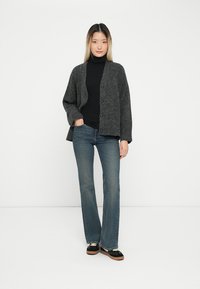 Grau Wollcardigan mit geknöpftem Vorderteil, getragen über einem schwarzen Rollkragenpullover und blauen Schlagjeans, kombiniert mit schwarzen Schuhen.