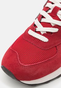 Punainen lenkkari, joka koostuu mokkasta ja verkkomateriaalista, valkoiset nauhat, pehmustettu nilkkareuna ja kumipohja.