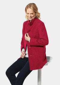 Fuchsia Wollmantel mit Stehkragen, durchgehender Knopfschließung und Seitennähten, getragen über einem hellen Pullover und dunklen Jeans.