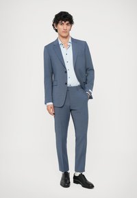 Fato azul claro com lapela recortada, casaco de um só botão e calças ajustadas, combinado com uma camisa azul claro de botões e sapatos pretos.