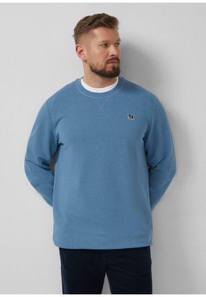 Mężczyzna z brodą w jasnoniebieskiej bluzie i ciemnych spodniach stoi na tle jasnoszarej ściany, patrząc w prawo, z rękami za plecami.