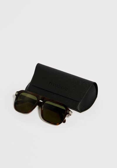 Lunettes de soleil écaille avec des verres foncés posées devant un étui à lunettes rectangulaire noir texturé sur une surface blanche.