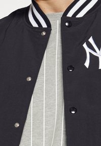 Svart varsityjacka med randig vit krage, utrustad med tryckknappar och en framträdande vit broderad logotyp på vänster sida över en grårandig skjorta.