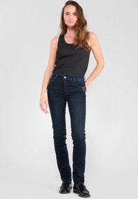 Jeans ajustados de color azul oscuro con una textura suave, que cuentan con una cintura de altura media, cinco bolsillos y herrajes plateados. Combinados con una camiseta de tirantes negra.