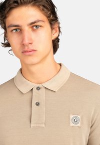 Camisa polo beige hecha de tejido texturizado que presenta un cuello con solapa, dos botones negros y un pequeño parche con el logo en el lado izquierdo del pecho.