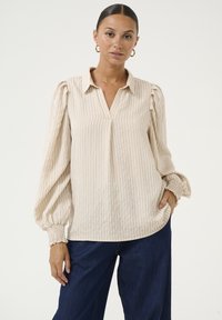 Beige gestreepte blouse met een kraag, v-hals en ingezette pofmouwen. Zachte, lichte stof met elastische manchetten, gecombineerd met donkerblauwe broeken.