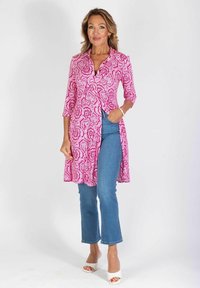 Różowa tunika w wzór paisley z zapięciem na guziki, rękawami 3/4 i bocznymi rozcięciami, zestawiona z jasnoniebieskimi rozkloszowanymi dżinsami i białymi szpilkami.