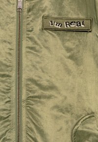 Žalios alyvuogių spalvos striukė su užtrauktuku, tekstūruota medžiaga ir pleistras, ant kurio parašyta "Aš esu tikras." Turi šonines kišenes su apvaliais kraštais.