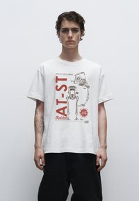 Bílý bavlněný tričko s grafickým potiskem robota AT-ST a červenými nápisy a akcenty. Krátké rukávy, volný střih, viditelné tetování na paži.