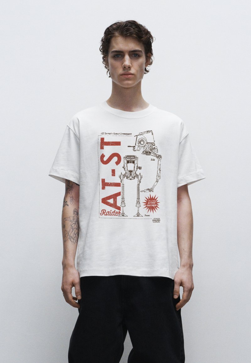 Bílý bavlněný tričko s grafickým potiskem robota AT-ST a červenými nápisy a akcenty. Krátké rukávy, volný střih, viditelné tetování na paži.