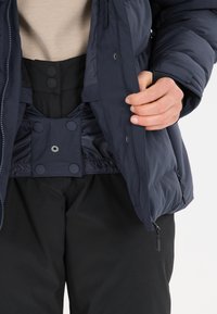 Marineblauer Steppjacke mit einem Reißverschluss vorne, Druckknöpfen und einem glatten Innenfutter, kombiniert mit schwarzen Hosen, die ein strukturiertes Design zeigen.