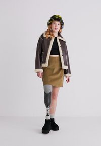 Brun shearling-fôret jakke, svart crop top, olivengrønn skjørt, svarte platå støvler og en fargerik pelshatt. Synlig proteseben.