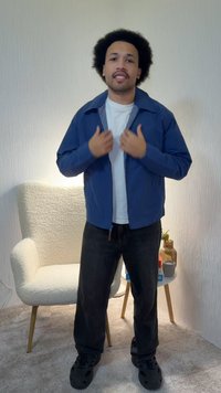 Άνδρας με σγουρά μαλλιά, φορώντας μπλε μπουφάν, λευκό μπλουζάκι και μαύρο παντελόνι, στέκεται σε ένα δωμάτιο με μια λευκή πολυθρόνα και ένα μικρό τραπέζι που κρατά ένα βάζο και μια κούπα.