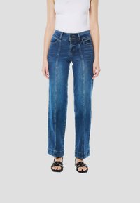 Blauwe denim wijde pijpen jeans met voorste naden, een hoge tailleband en contrasterende stiksels. Gecombineerd met zwarte sandalen met studs.