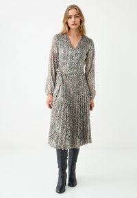 Vrouw in een lange mouw leopardenprint midi-jurk met een tailleband en zwarte hoge laarzen met hakken, staand tegen een effen witte achtergrond.