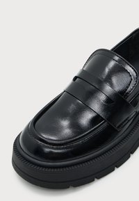 Gros plan d'un mocassin en cuir noir brillant avec une semelle épaisse à rainures et une bride sur le dessus à l'avant.