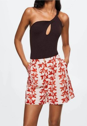 Vrouw die een mouwloos zwart asymmetrisch topje draagt met een keyhole aan de voorkant en een witte short met rode bloemenprint, handen in de zakken.