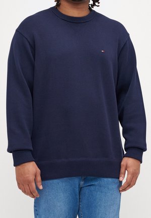 Personne portant un pull en maille bleu marine avec poignets et ourlet côtelés, assorti d’un jean bleu clair, sur un fond uni.
