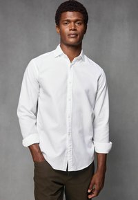 Weißes Button-Down-Hemd mit langen Ärmeln und umgeschlagenen Manschetten, aus glattem Stoff, kombiniert mit dunkelbraunen Chinos. Einfaches, modernes Design.