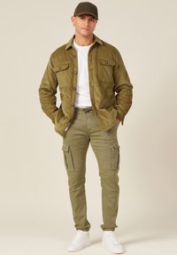Olivegrüne Cordjacke mit vier Taschen, getragen über einem weißen T-Shirt und olivfarbenen Cargo-Hosen. Weiße Sneaker vervollständigen das Outfit.