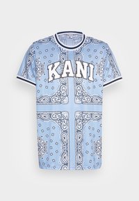 Světle modré tričko s krátkým rukávem s černobílým paisley vzorem, které má na přední straně výrazné logo "KANI" a pruhovaný límec.