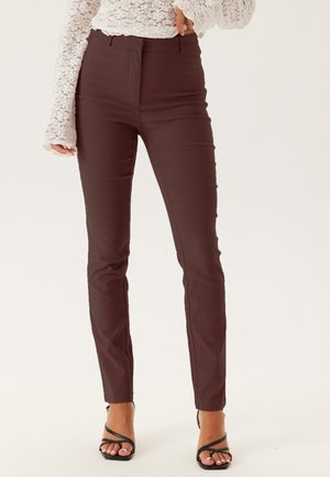Pantalon marron slim-fit porté avec un haut blanc en dentelle à manches longues et des sandales à talons hauts noires à brides sur fond blanc.
