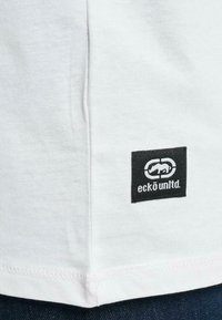 Witte katoenen t-shirt met een zwart gestikt etiket met een logo en de tekst "eckō unltd." aan de onderlinkse kant nabij de zoom.
