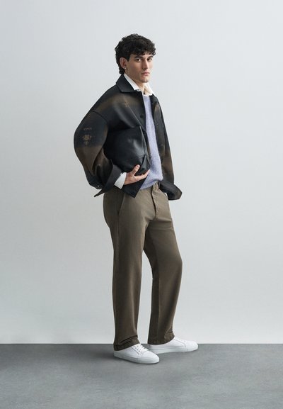 Μαύρο δερμάτινο μπουφάν με ξεθωριασμένο σχέδιο, ανοιχτό μωβ πλεκτό πουλόβερ, καφέ παντελόνι και λευκά αθλητικά παπούτσια. Το μοντέλο κρατά μια μαύρη τσάντα.