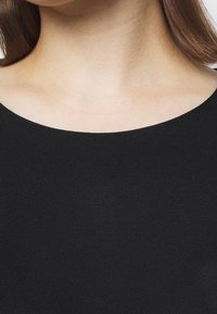 Svart tyg med en slät textur, med en bred, rund halsringning. Designen är enkel och kroppsnära, utan utsmyckningar.