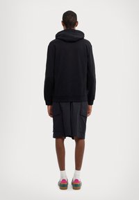 Svart hettegenser, kombinert med mørke cargo-shorts med lommer. Fottøyet inkluderer flerfargede klip-klapper og hvite sokker.
