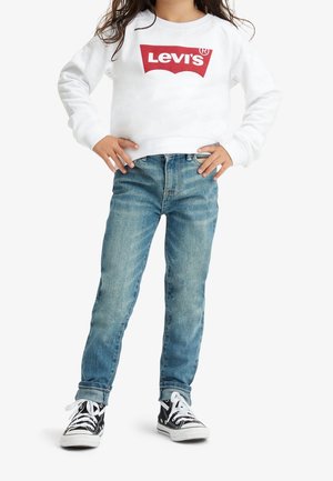 Kind draagt een wit Levi's sweatshirt, opgerolde blauwe spijkerbroek en zwarte hoge sneakers met witte veters, staat met de handen in de zij.