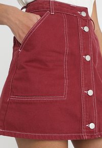 Jupe en denim rouge avec des coutures blanches, poche latérale et fermeture à boutons à l'avant. Forme structurée et tissu texturé.