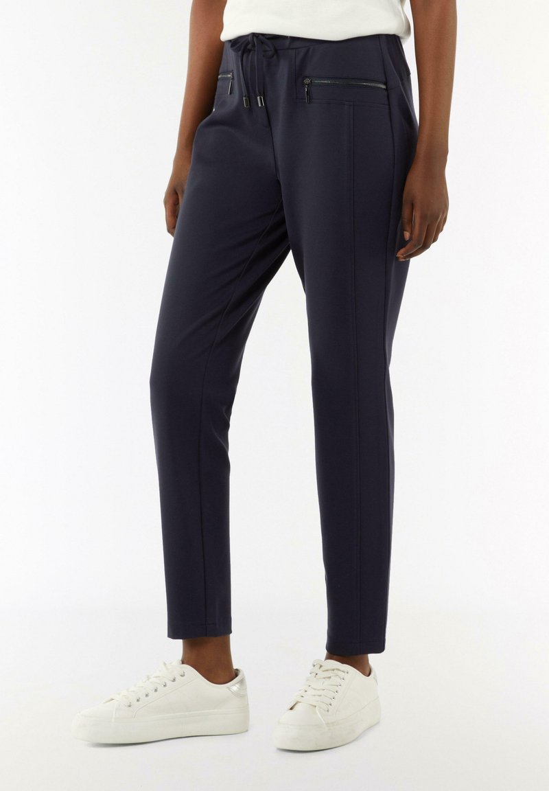 Navy stretchbroek met een tailleband met trekkoord, voorzakken met rits en platte naad aan de voorkant. Gecombineerd met witte sneakers.