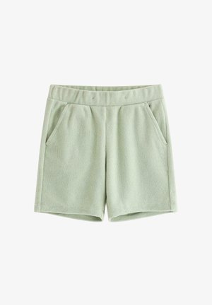 Smilende ung dreng iført lysegrøn polo og shorts, beige sokker med grønne striber og hvide sneakers, stående på ensfarvet baggrund.