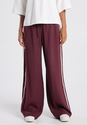 Bordeauxkleurige wijde broek van zachte stof, met crèmekleurige zijstrepen, een elastische tailleband en een relaxed fit. Gepaard met witte schoenen.