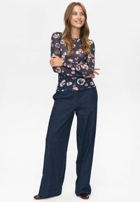 Donna con un sorriso, capelli lunghi, che indossa una blusa a maniche lunghe floreale blu marino, pantaloni larghi blu scuro e scarpe scure, in piedi con le braccia incrociate.